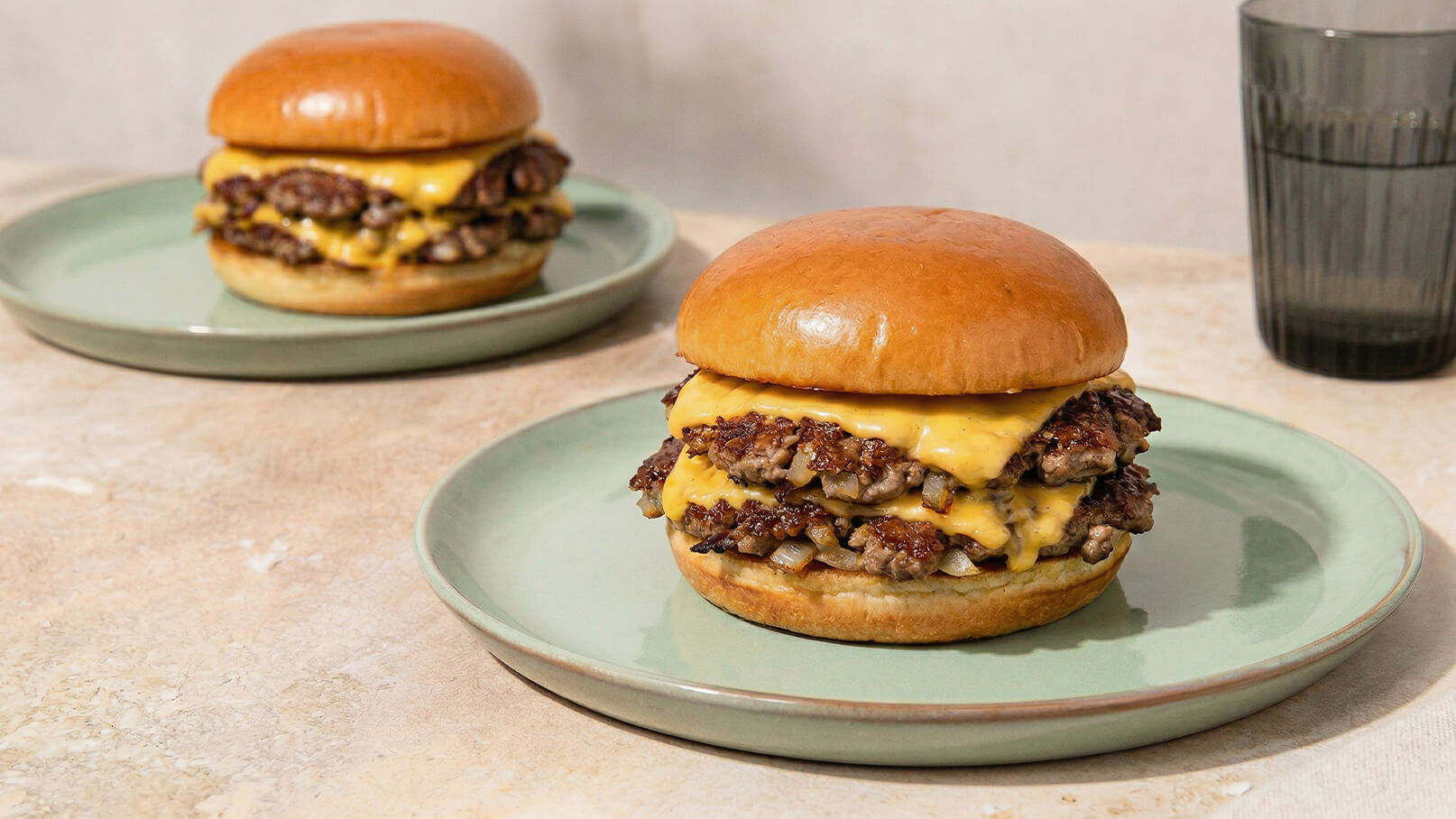 Zwei Hamburger mit geschmolzenem Cheddar-Käse auf Brötchen liegen auf Tellern. Der Vordergrund zeigt einen Hamburger mit zwei Fleischpattys und Käse. Im Hintergrund steht ein weiteres ähnliches Gericht sowie ein Glas Wasser.
