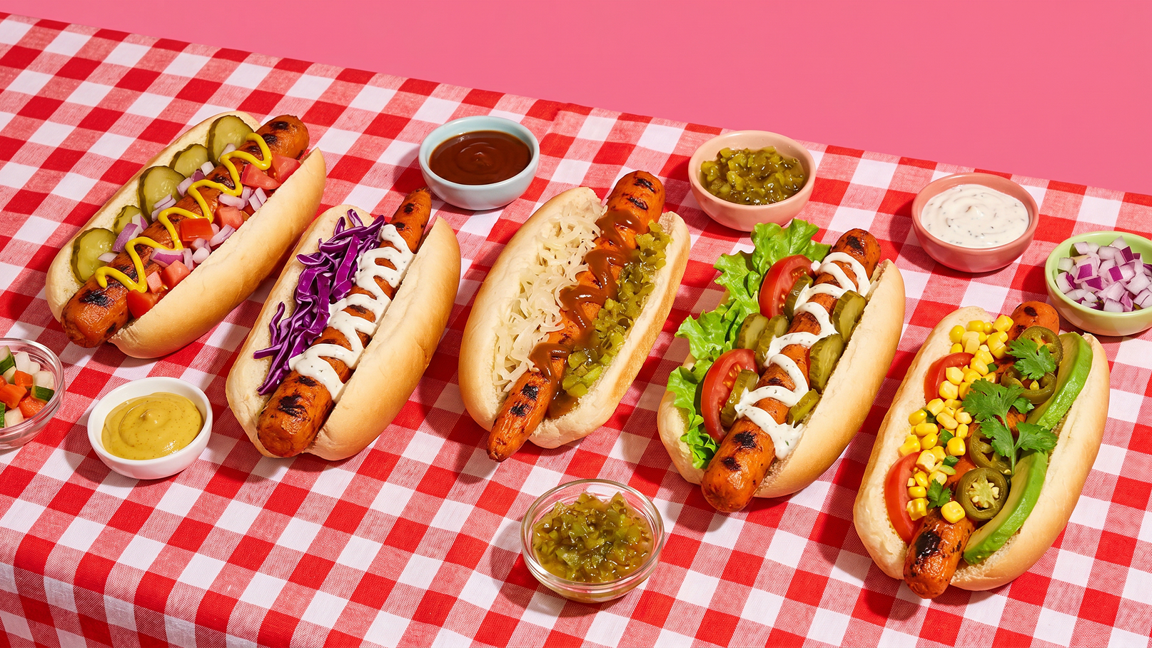 Sechs unterschiedlich belegte Hotdogs (Würstchen im Brötchen mit Senf, Ketchup, Krautsalat, Essiggurken, Jalapeños, Mais und Salat) auf rot-weiß kariertem Tischtuch, mehrere kleine Schälchen mit Saucen daneben.