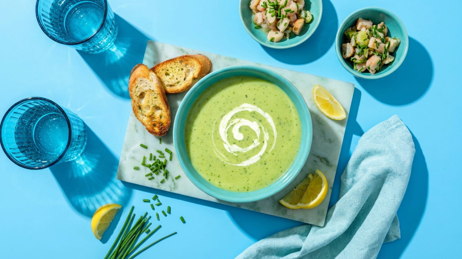 Eine grüne Suppe mit Sahnewirbel in einer blauen Schüssel, serviert auf einem weißen Unterlage. Daneben befinden sich geröstete Brotscheiben, Zitronenscheiben und eine kleine Schale mit Salat. Im Hintergrund stehen zwei blaue Gläser.