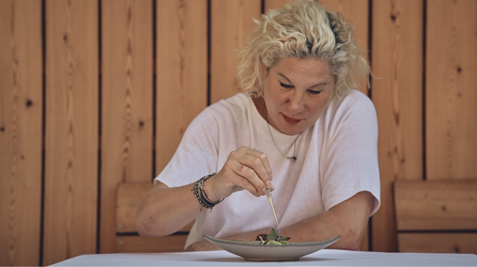 Eine Frau mit lockigem, blondem Haar sitzt an einem Tisch und arbeitet konzentriert mit einer Pinzette. Vor ihr befindet sich ein Teller mit kunstvoll angerichtetem Essen. Der Hintergrund besteht aus Holzpaneelen, die eine warme Atmosphäre schaffen.