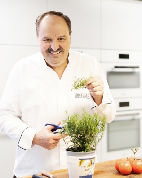 Ein lächelnder Koch in einer weißen Kochjacke schneidet frische Rosmarinzweige aus einem Blumentopf. Auf dem Tisch liegen zwei Tomaten. Der Hintergrund zeigt eine moderne Küche.
