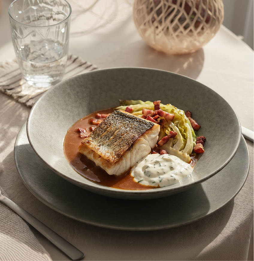 Gebratenes Fischfilet mit knuspriger Haut auf brauner Sauce, dazu gedünsteter Wirsing mit Speckwürfeln und ein Klecks Kräutercreme in einem tiefen Teller.