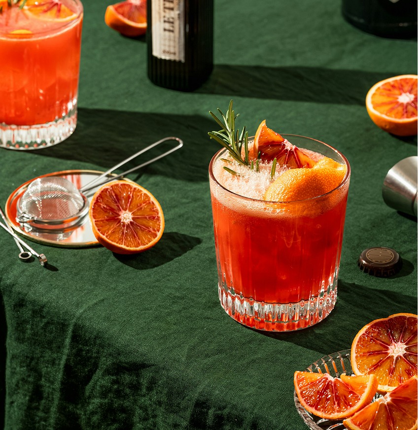 Nahaufnahme eines Cocktailglases mit blutrotem Blutorangensaft, garniert mit Rosmarin und Orangenzeste, umgeben von halbierten und aufgeschnittenen Blutorangen und Barwerkzeug auf dunkelgrünem Tuch.