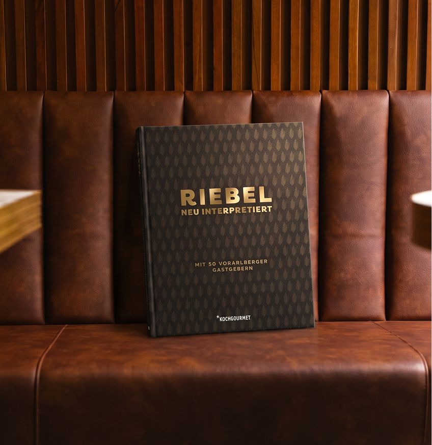Ein gebundenes, schwarzes Buch mit goldener Prägung "RIEBEL NEU INTERPRETIERT" lehnt auf einer braunen Lederbank vor Holzpaneelen.