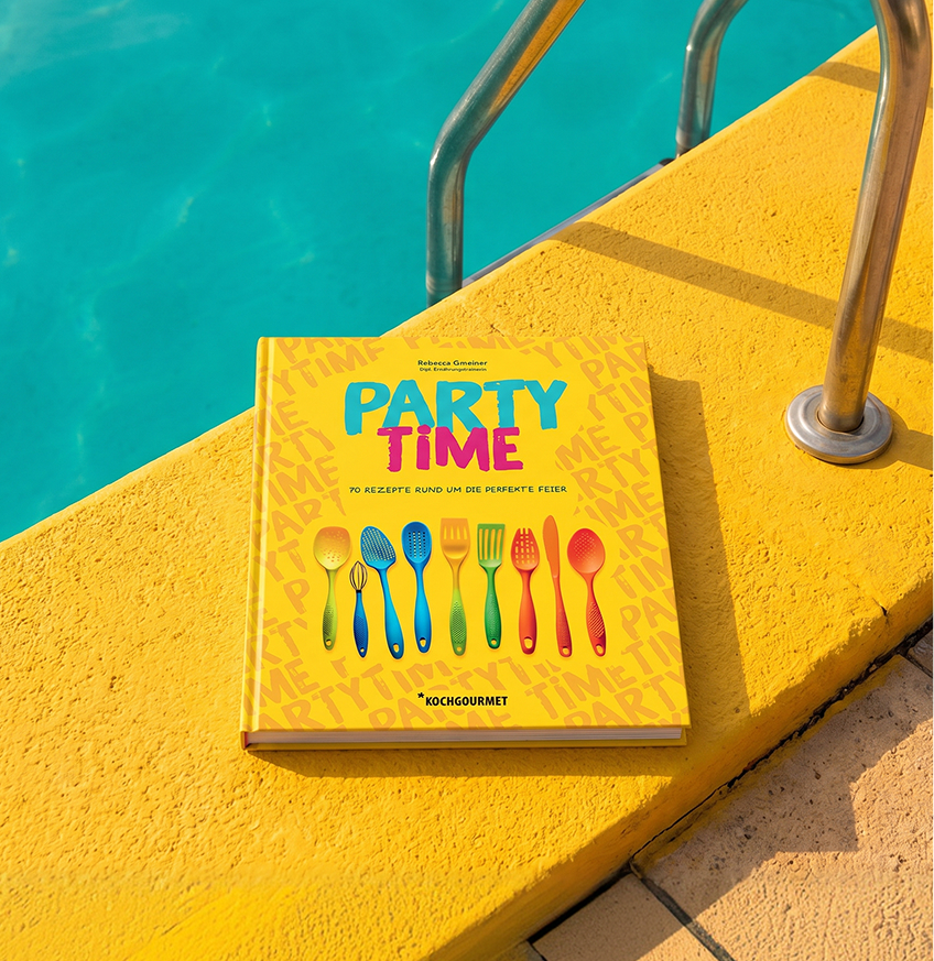 Ein buntes Kochbuch mit Titel „PARTY TIME“ und Abbildungen farbiger Löffel und Gabeln liegt auf dem gelben Beckenrand neben türkisblauem Poolwasser.