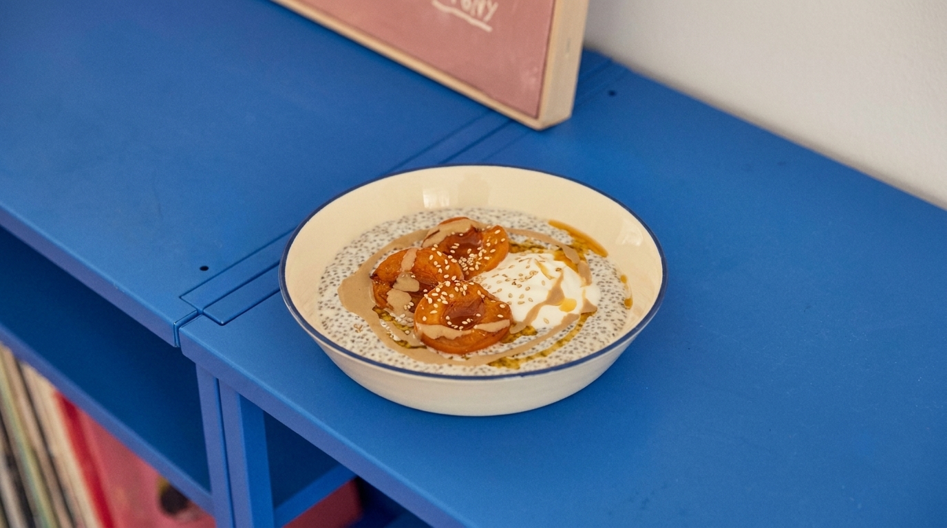 Eine weiße Schüssel steht auf einem blauen Tisch. In der Schüssel befinden sich Chia-Pudding, eingelegte Früchte, Joghurt und ein Honig- oder Sirupüberzug. Das Gericht ist einfach, aber ansprechend angerichtet.