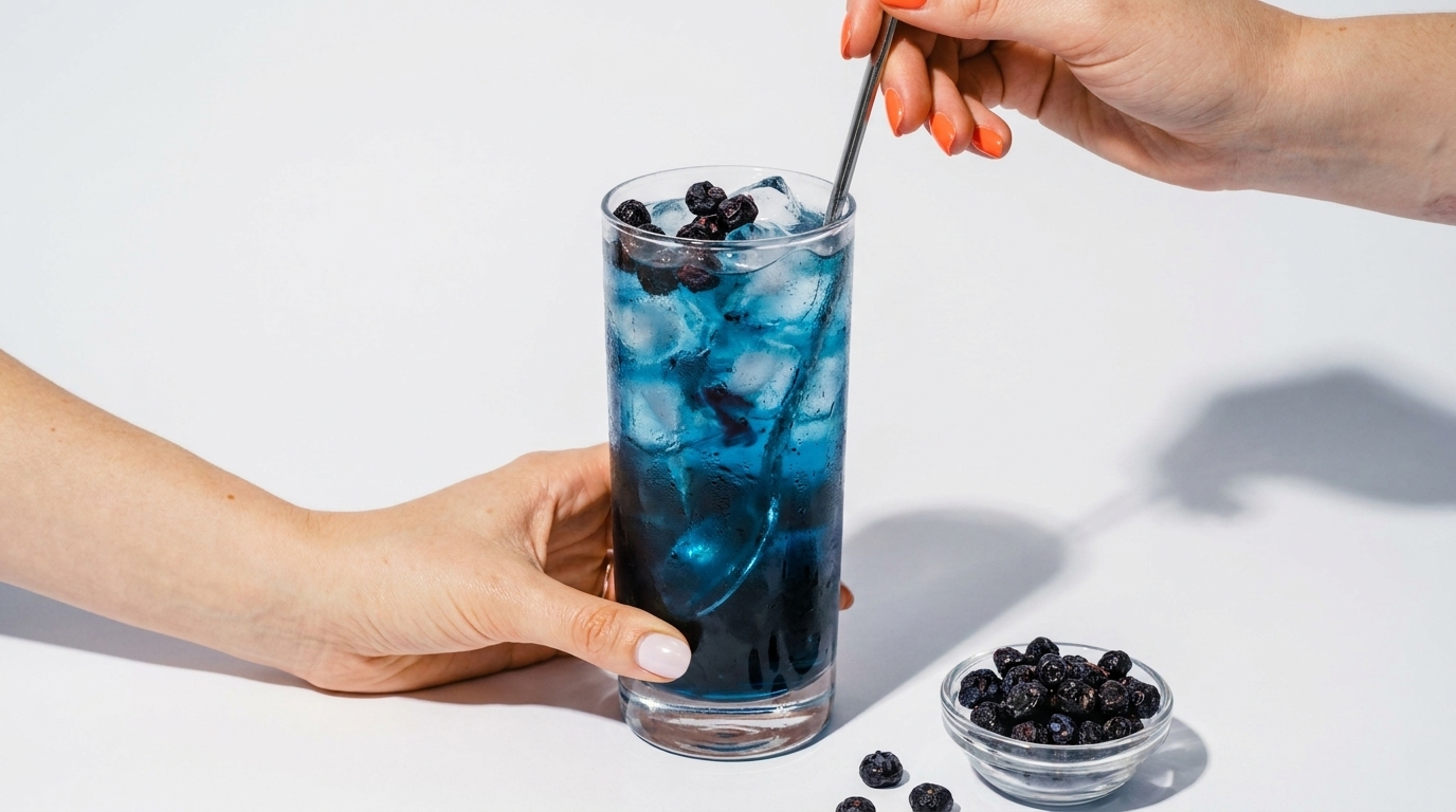 Zwei Hände bereiten ein blaues Eisgetränk in einem hohen Glas: eine Hand hält das Glas, die andere rührt mit einem Löffel. Eiswürfel und Blaubeeren im Getränk; daneben eine kleine Schale und einige lose Blaubeeren.