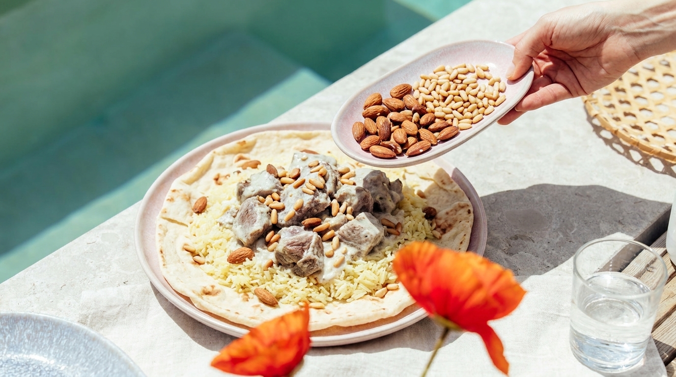 Teller mit Fladenbrot, Reis und gewürzten Fleischstücken, bestreut mit Mandeln und Pinienkernen; eine Hand streut gerade Nüsse darüber.