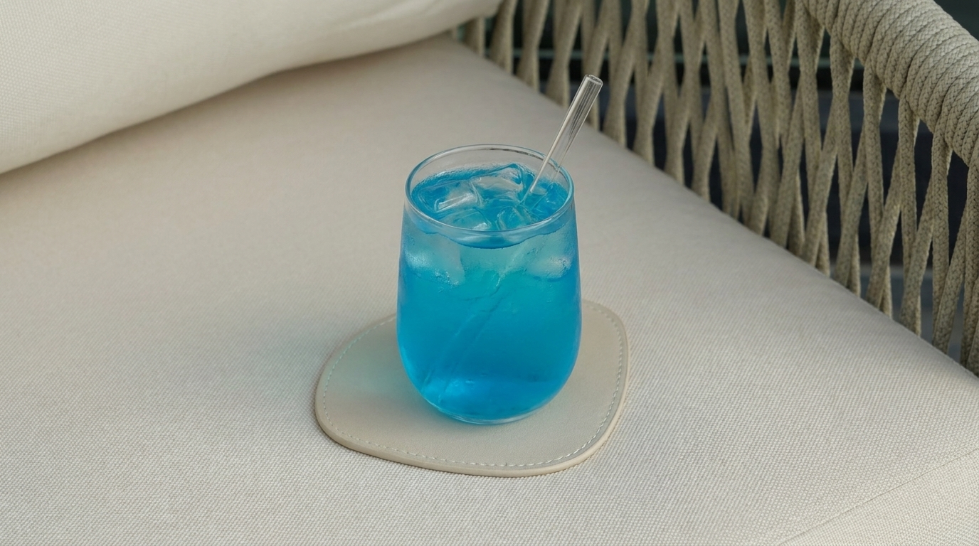 Glas mit leuchtend blauem Getränk, Eiswürfeln und Strohhalm auf einem hellen Untersetzer, abgestellt auf einem hellen Sofa.
