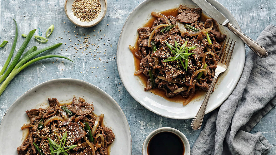 Zwei Teller mit asiatischem Rindfleisch-Gericht, garniert mit Frühlingszwiebeln und Sesam. Der Hauptteller zeigt zarte Stücke Fleisch in einer dunklen Sauce, umgeben von Zwiebeln und grünen Gemüsestreifen. Eine kleine Schüssel mit Sauce und einige Sesamsamen sind ebenfalls zu sehen.