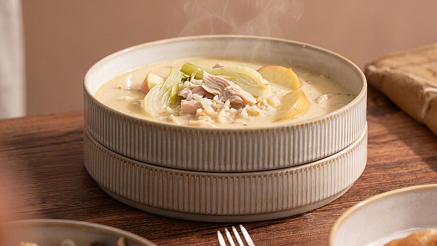 Ein tiefes Schüsselgericht mit dampfender, cremiger Suppe, das mit Hühnchenstücken, Sellerie und kleinen Kartoffeln garniert ist. Die Suppe wird auf einem rustikalen Holztisch serviert, umgeben von einem weiteren Teller und einem Tuch.
