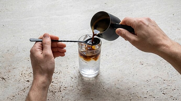 Zwei Hände gießen Espresso aus einem Kännchen über einen Barlöffel in ein Glas mit Eis und Tonic Water.