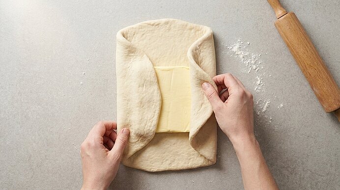 Zwei Hände falten einen Teigmantel um einen rechteckigen Butterblock, typischer Arbeitsschritt zur Herstellung von Blätterteig.