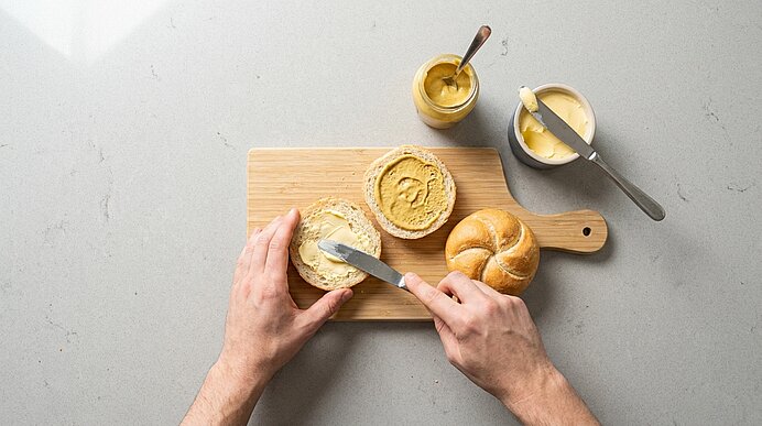 Zwei Hände bestreichen eine Semmelhälfte auf einem Holzbrett mit Butter. Daneben liegt eine mit Senf bestrichene Hälfte.