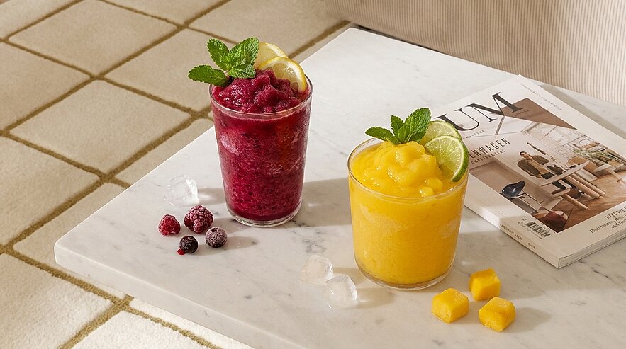 Zwei Gläser Fruchtslushies auf einer Marmorplatte: ein dunkelroter Himbeer-Slush mit Minze und Zitronenscheibe, daneben ein gelber Mangoslush mit Minze und Limettenscheibe; Himbeeren, gefrorene Beeren und Mangowürfel liegen daneben.