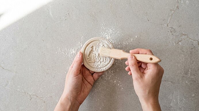 Zwei Hände halten eine runde Gipsform mit feinem Relief; mit einem kleinen Holzspatel wird überschüssiger Gipsstaub entfernt.