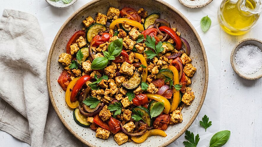 Eine Schüssel mit buntem Gemüse und Tofu, darunter rote, gelbe und grüne Paprika, Zucchini und Kirschtomaten. Frische Kräuter wie Basilikum und Petersilie sind dekorativ darüber gestreut. Die Speise sieht appetitlich und gesund aus.