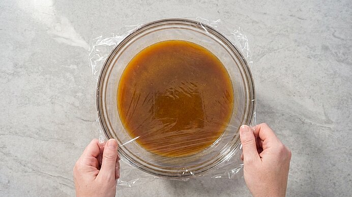 Zwei Hände ziehen Frischhaltefolie über eine Glasschüssel mit brauner Sauce auf einer hellen Arbeitsfläche.