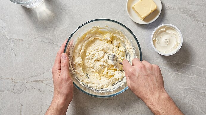 Zwei Hände rühren mit einem Löffel eine cremige Butter‑/Frischkäse‑Mischung in einer Glasschüssel; ein Stück Butter und ein Frischkäsebehälter stehen daneben.