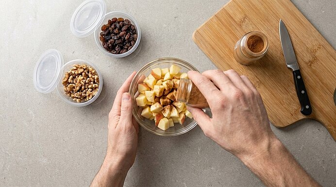 Hände streuen Zimt über gewürfelte Äpfel in einer Glasschüssel; daneben Schalen mit Rosinen und Walnüssen sowie ein Schneidebrett mit Messer.
