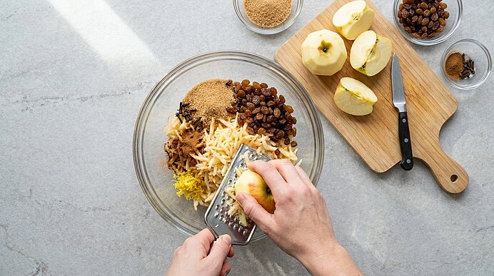 Hände reiben einen geschälten Apfel mit einer Reibe über eine Glasschüssel, in der geriebener Apfel, brauner Zucker und Rosinen liegen; daneben ein Schneidebrett mit halbierten Äpfeln und Messer.