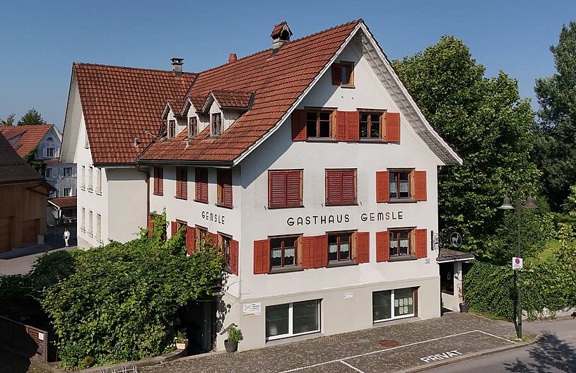 Das Bild zeigt das Gasthaus Gemsle, ein weißes Gebäude mit roten Fensterläden und einem Satteldach. Der Schriftzug "GASTHAUS GEMSL" ist gut sichtbar. Umgeben ist das Haus von Bäumen und Grünflächen, bei klarem blauen Himmel.