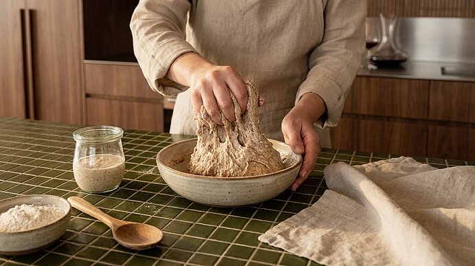 Hände kneten Brot­teig in einer Schüssel auf der Arbeitsfläche; daneben Glas mit Sauerteig, Schüssel mit Mehl und ein Holzlöffel.