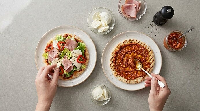 Zwei Hände bestreichen Pitabrot mit Pesto und belegen eine Salatpizza mit Mozzarella, Schinken, Paprika und Salat.