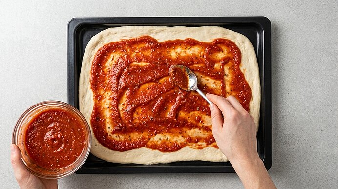 Zwei Hände verstreichen mit einem Löffel Tomatensauce aus einer Schüssel auf rohem Pizzateig auf einem Backblech.