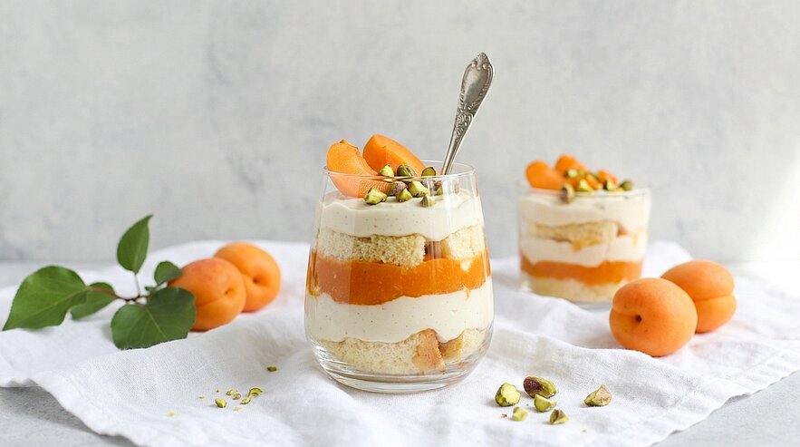 Zwei Gläser mit Schichtdessert aus Aprikosen, heller Creme und Biskuitstücken, oben mit Aprikosenstücken und gehackten Pistazien garniert; frische Aprikosen daneben.