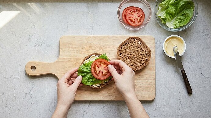 Zwei Hände belegen die untere Brötchenhälfte auf einem Schneidebrett mit Salat und einer Tomatenscheibe.