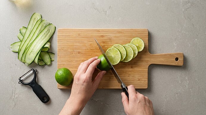 Zwei Hände schneiden eine Limette auf einem Holzbrett in Scheiben. Daneben liegen Gurkenbänder und ein Sparschäler.