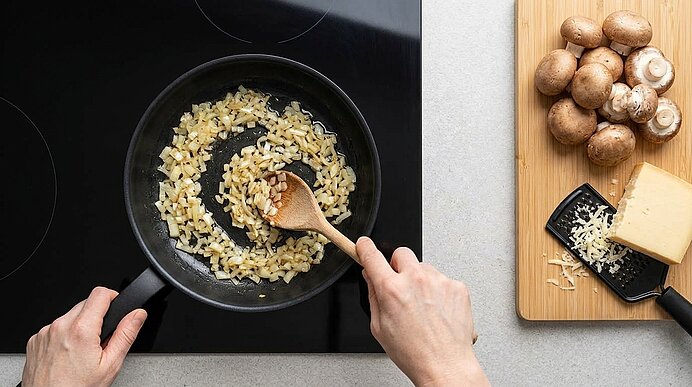 Zwei Hände rühren glasige Zwiebelwürfel in einer Pfanne. Rechts liegen Champignons und Bergkäse mit einer Reibe.