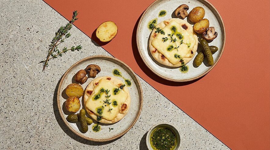Zwei Teller mit geschmolzenem Raclettekäse auf Kartoffeln, dazu gebratene Champignons, Cornichons und Kräuteröl, mit frischem Thymian garniert.