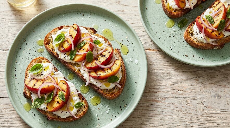 Mehrere geröstete Brotscheiben belegt mit cremigem Frischkäse, Pfirsich- oder Nektarinenstücken, dünnen roten Zwiebelringen, frischen Basilikumblättern und Olivenöl auf grünen Tellern.