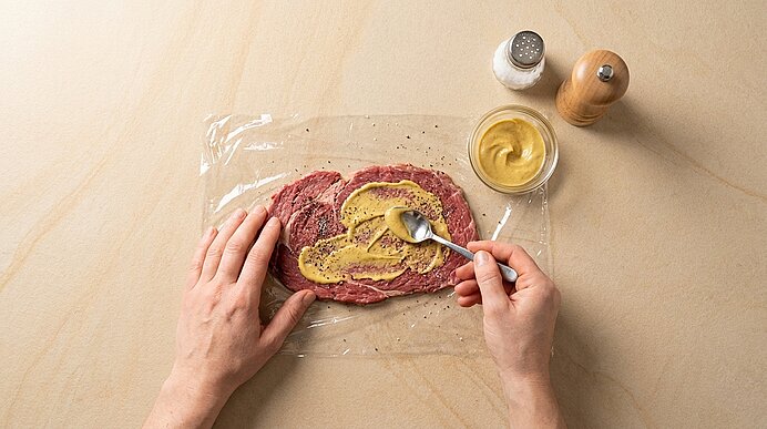 Zwei Hände bestreichen rohes, dünn geschnittenes Rindfleisch auf Frischhaltefolie mit Senf; Salz- und Pfefferstreuer sowie ein Senfglas daneben.