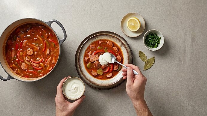 Schüssel mit roter Gulaschsuppe mit Wurstscheiben, Kartoffeln und Gemüse; Hand gibt Löffel mit Sauerrahm dazu. Daneben Topf, Zitronenhälfte und Schale mit gehackten Kräutern.