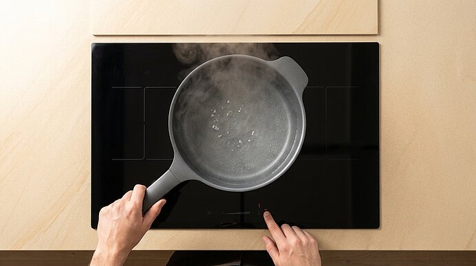 Draufsicht: Hände halten einen dampfenden Topf auf einem schwarzen Induktionskochfeld; eine Hand berührt das Touch-Bedienfeld.