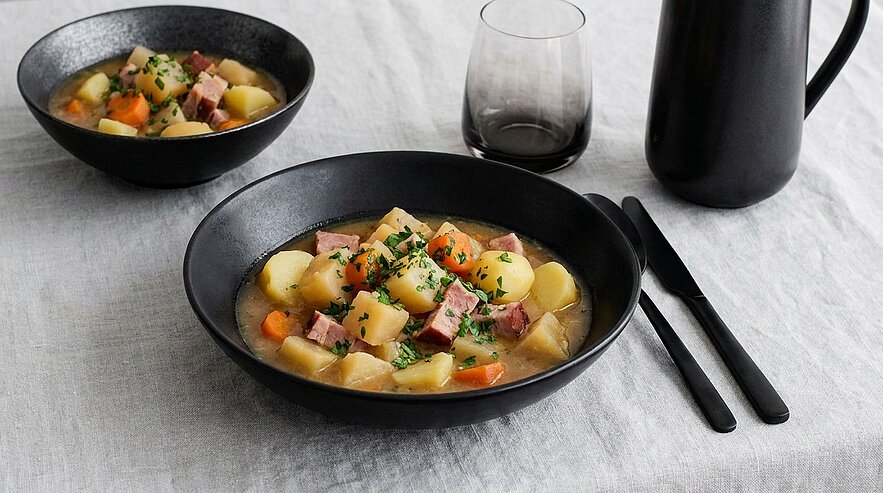 Zwei schwarze Schalen mit herzhaftem Eintopf aus Kartoffeln, Karotten und Schinken, garniert mit Petersilie; daneben schwarzes Besteck, ein Glas und eine Kanne.