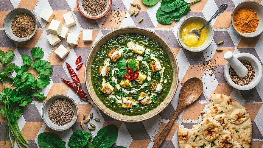 Eine Schüssel mit grünem Spinat-Curry, garniert mit Würfeln von gebratenem Käse, frischen Kräutern und einem hübschen Muster aus Joghurt und rote Chilischoten. Daneben sind verschiedene Gewürze, frisches Gemüse und Stücke von Fladenbrot arrangiert.