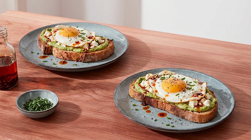 Zwei Teller mit Avocado-Toast: dicke Brotscheiben bestrichen mit zerdrückter Avocado, je ein Spiegelei obenauf, gehackter Schnittlauch und Spritzer würziger Sauce; daneben kleine Schale mit zusätzlichem Schnittlauch.