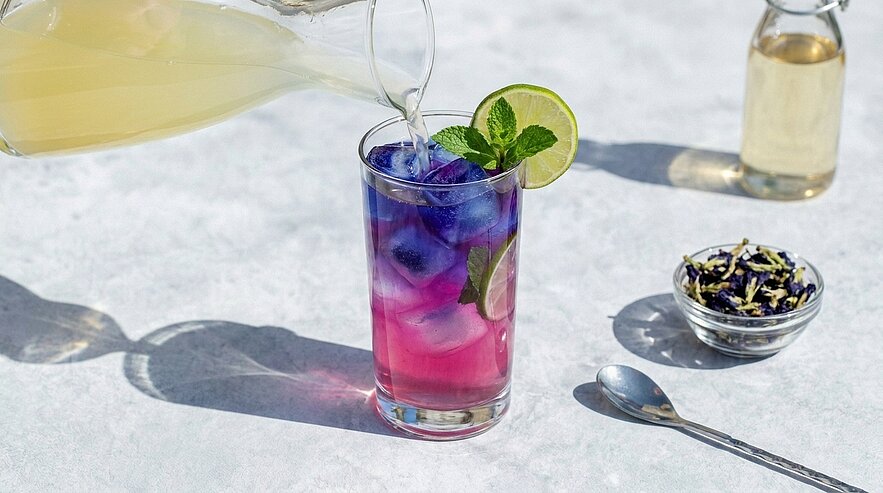 Ein hohes Glas mit farbigem Schichtgetränk (violett zu pink) über Eis, garniert mit Limettenscheibe und Minzblatt, während aus einem Krug eine helle Flüssigkeit eingeschenkt wird.