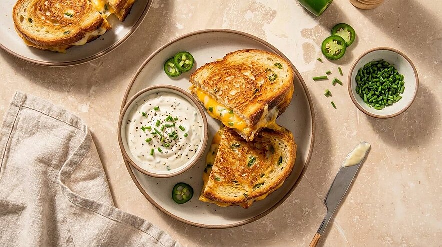 Zwei gegrillte Käsesandwiches mit verlaufendem geschmolzenem Käse auf einem Teller, dazu eine Schüssel mit cremigem Dip, gehacktem Schnittlauch und Jalapeño-Scheiben.