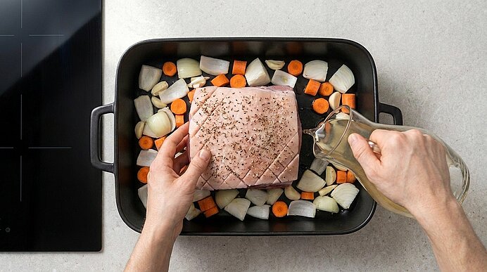 Zwei Hände legen einen gewürzten Braten in einen Bräter auf ein Bett aus geschnittenen Karotten und Zwiebeln; mit der anderen Hand wird Öl über das Fleisch gegossen.