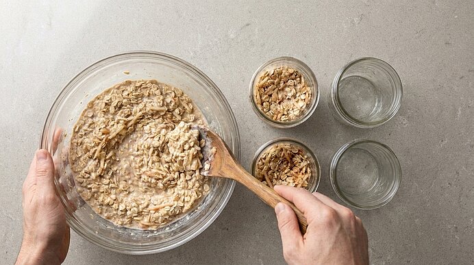 Zwei Hände rühren mit einem Holzlöffel eine Glasschüssel mit Haferflocken-Mischung; daneben drei kleine Gläser mit Haferflocken und Wasser.