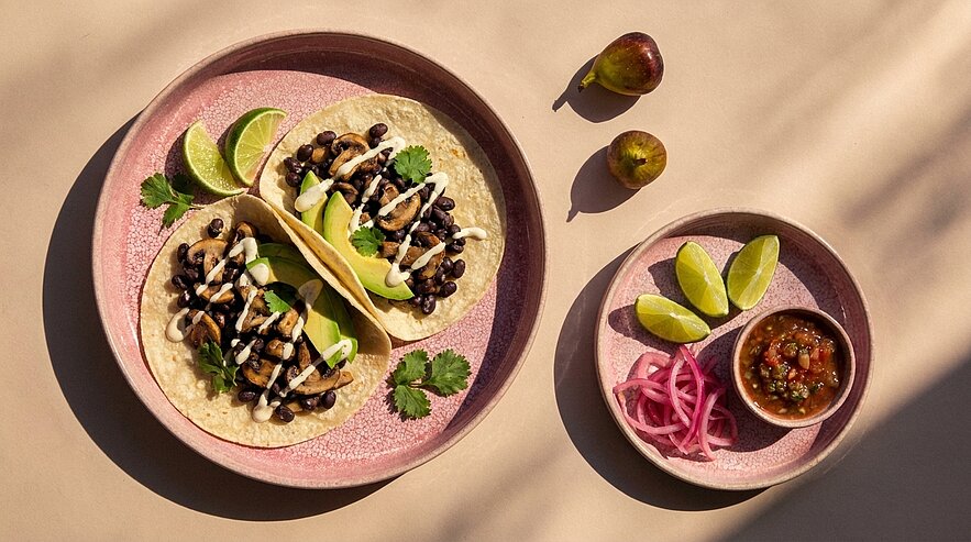 Zwei Tortilla-Tacos gefüllt mit schwarzen Bohnen, gegrilltem Gemüse, Avocadoscheiben und weißer Sauce, serviert mit Limetten, eingelegten roten Zwiebeln und Salsa.
