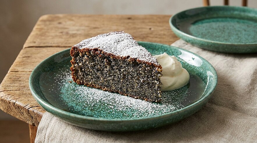 Auf einem grünen Teller liegt ein Stück Mohnkuchen, bestäubt mit Puderzucker. Neben dem Kuchen befindet sich ein Klecks Sahne. Der Teller steht auf einem rustikalen Holztisch, der einen warmen, einladenden Eindruck vermittelt.