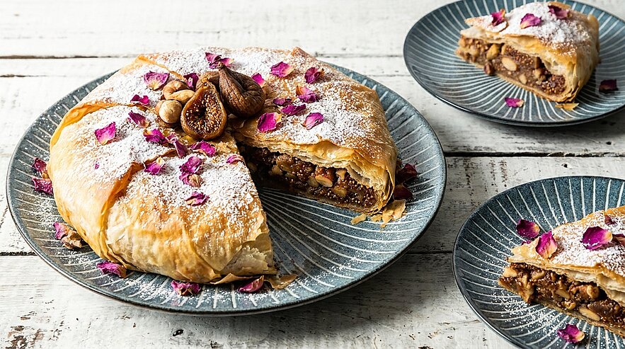 Ein frisch gebackenes, rundes Gebäck mit knusprigem Teig und einer Füllung aus Nüssen, Trockenfrüchten und Gewürzen. Das Gebäck ist mit Puderzucker und getrockneten Rosenblüten dekoriert. Auf einem separaten Teller sind Stücke des Gebäcks angerichtet.