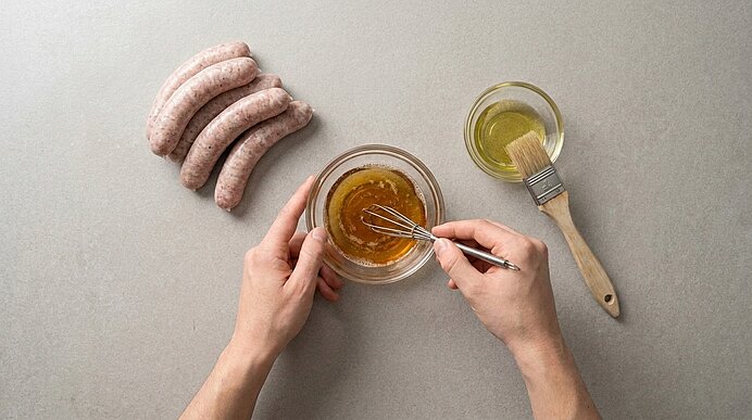 Zwei Hände rühren in einer Glasschüssel eine senfartige Marinade; daneben liegen vier rohe Bratwürste und eine Schale Öl mit Pinsel.