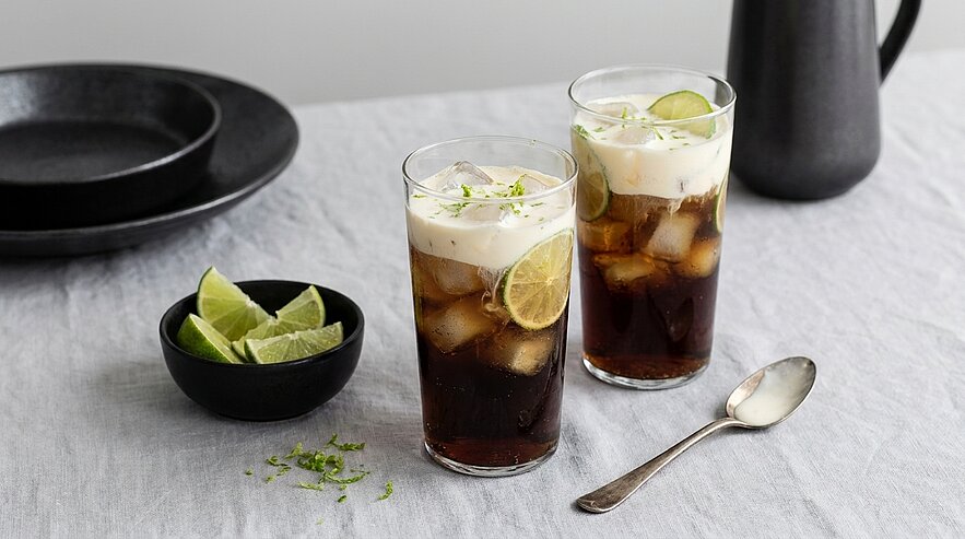Zwei hohe Gläser mit eisgekühltem Cola-Getränk, Schaumhaube und Limettenscheiben; daneben eine kleine Schale mit Limettenkeilen.